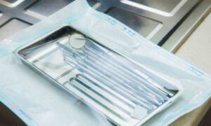 A selection of clean, metal dental instruments sits in a small tray. The tray is wrapped in sterile medical device packaging.