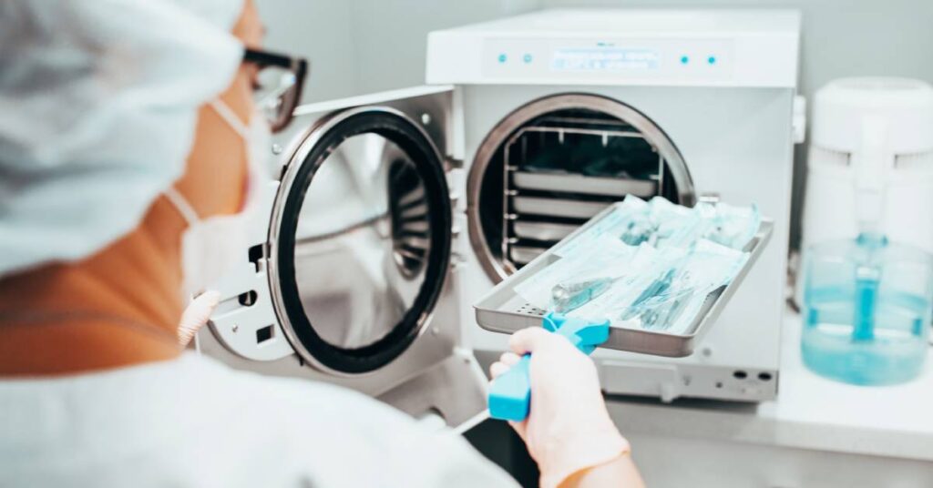 A woman wearing gloves and a mask loads a tray of medical instruments into a machine for sterilization.