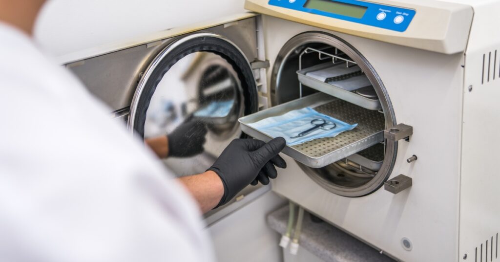 A medical professional wearing gloves places a tray of sealed medical devices into an autoclave for cleaning.