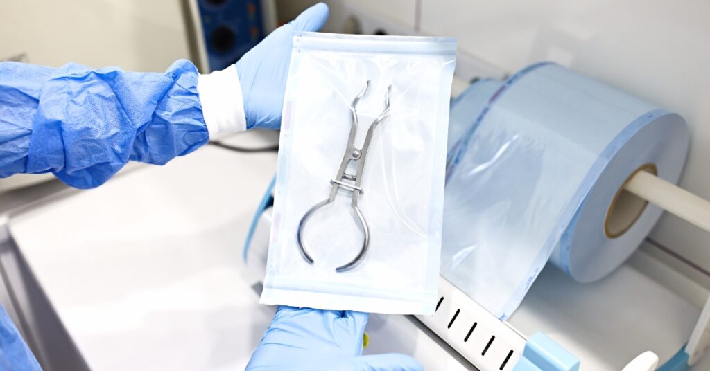 A healthcare professional wearing gloves holds up a pair of forceps in sealed, sterile medical packaging.