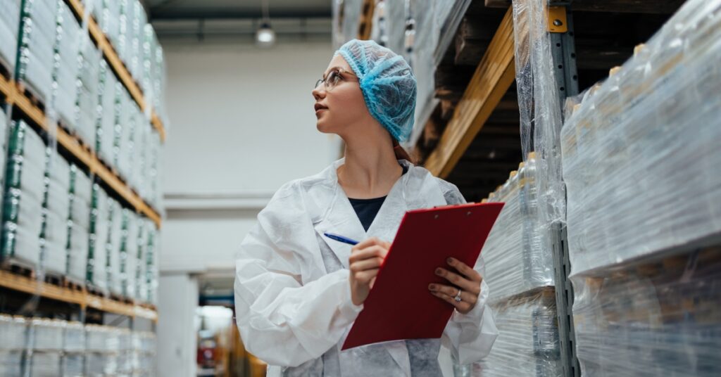 A woman wearing a lab coat and hair net inspects pallets of packaged products. She is holding a clipboard and pen.