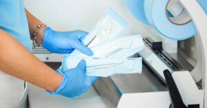 A person holds three dental instruments in secure, sterile wrapping. They are wearing bracelets and blue medical gloves.