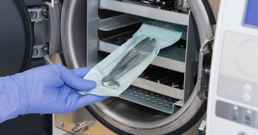 Someone placing a metal medical instrument wrapped in clear packaging onto the shelf of an autoclave.