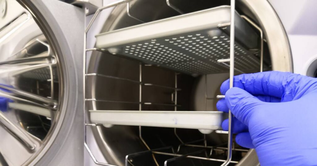 Someone pulling out a rack of metal trays from inside a steam sterilizer. They are wearing blue medical gloves.