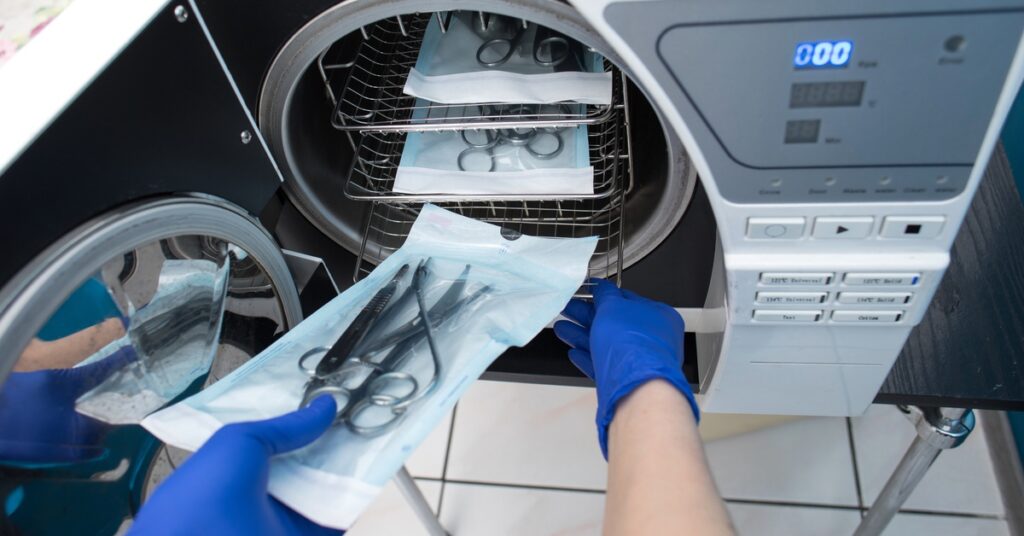 Someone wearing blue gloves while placing medical instruments wrapped in a sterilization pouch into an autoclave.