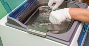 A medical professional wearing gloves places metal tools into the water-filled tray of an ultrasonic cleaner.