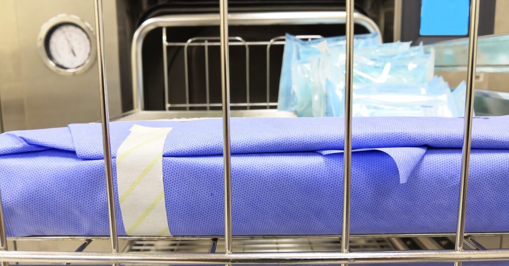 Blue sterilization wrap around a product sitting on a wire rack. There are sterilization pouches on the rack behind it.