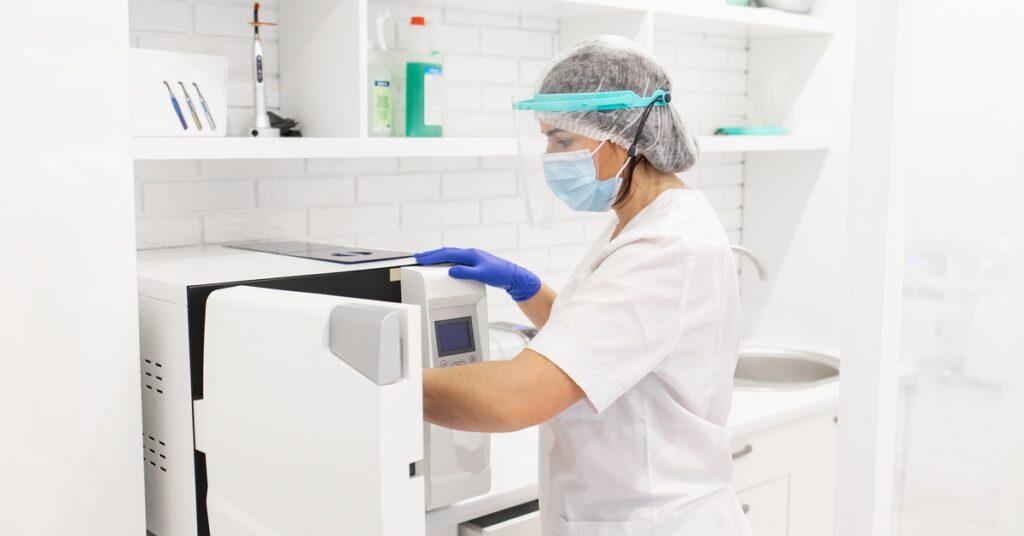 A worker wearing scrubs, a face shield, a face mask, disposable gloves, and a bouffant cap reaches into an autoclave.