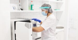 A worker wearing scrubs, a face shield, a face mask, disposable gloves, and a bouffant cap reaches into an autoclave.