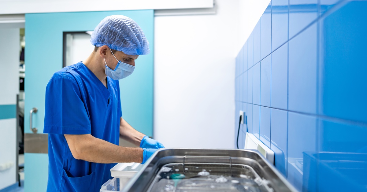 A man in blue scrubs, a hairnet, and a face mask is standing next to a wall and is reaching into a plastic box.