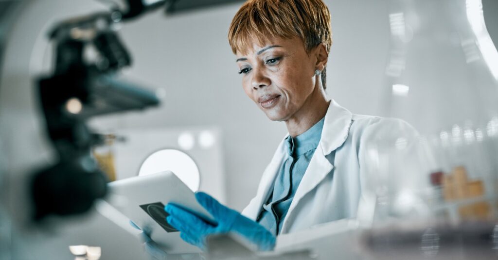 A woman in a laboratory, holding a tablet and surrounded by beakers, a microscope, and various other tools.