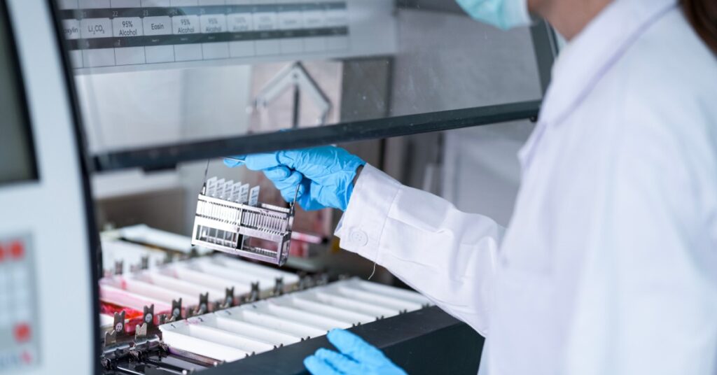 A lab technician wearing a mask and gloves loads a sample rack into a machine that has a transparent cover.