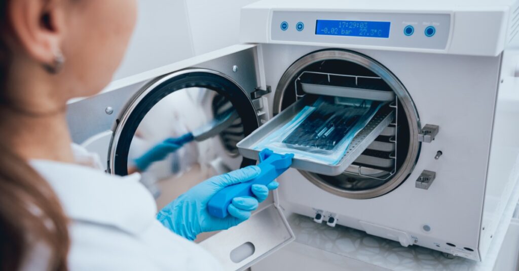 A technician wearing blue gloves uses a tray lifter to slide a tray of sealed instruments into an autoclave chamber.
