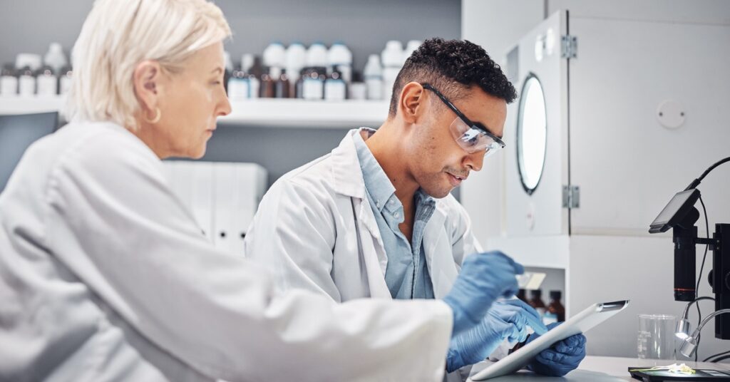 Two colleagues in white coats and blue gloves review information on a tablet. Bottles are behind them in the laboratory.