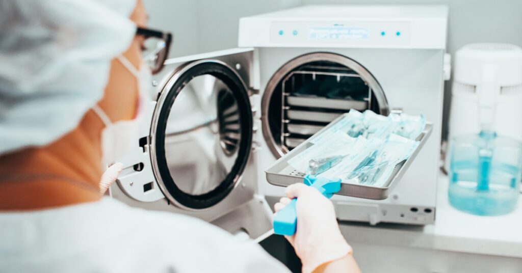 A lab worker wearing gloves, a mask, and a hair covering holds a tray of packaged instruments near a sterilization unit.