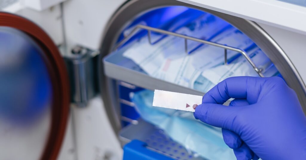 A gloved hand holds a sterilization indicator strip in front of a sterilizer chamber that holds wrapped medical items.