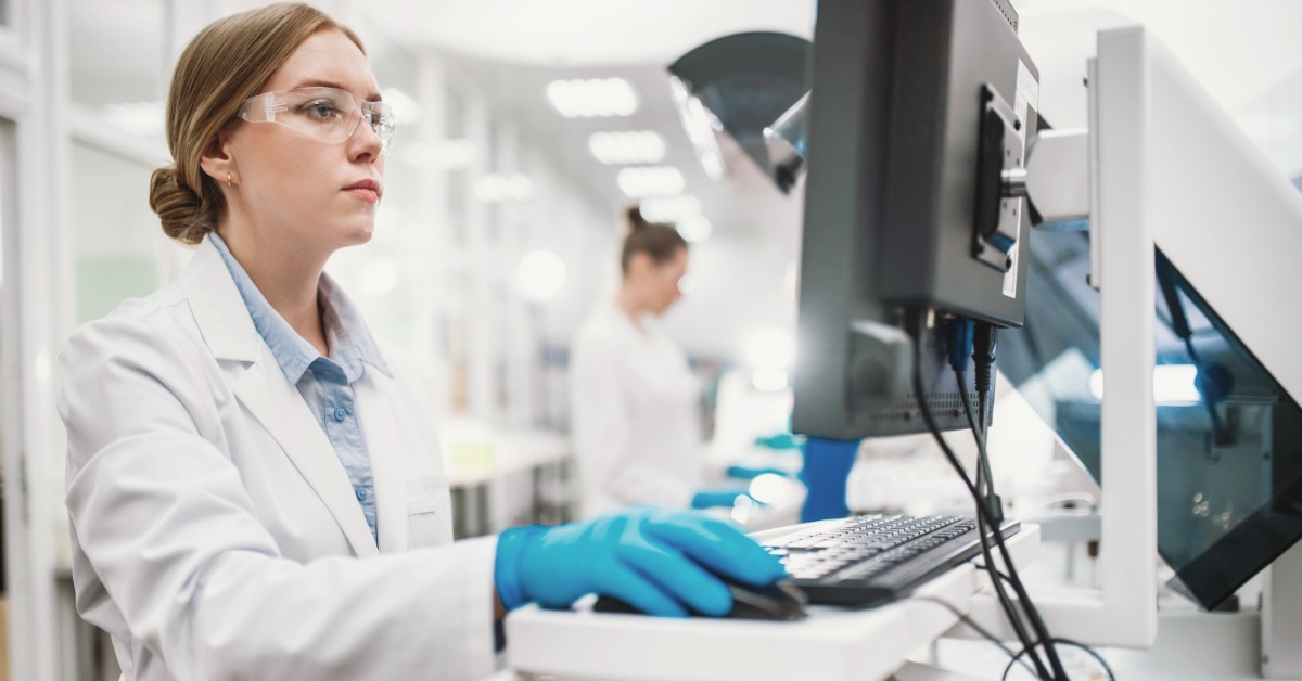 A worker in blue gloves, safety glasses, and a white lab coat looks at a screen and moves a mouse at a computer station.