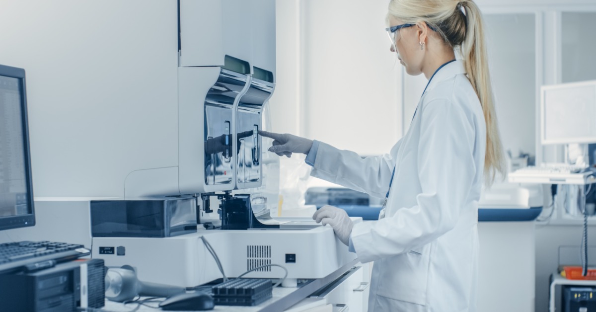 A lab worker in a white coat, gloves, and safety glasses presses a button on the screen of a large laboratory machine.