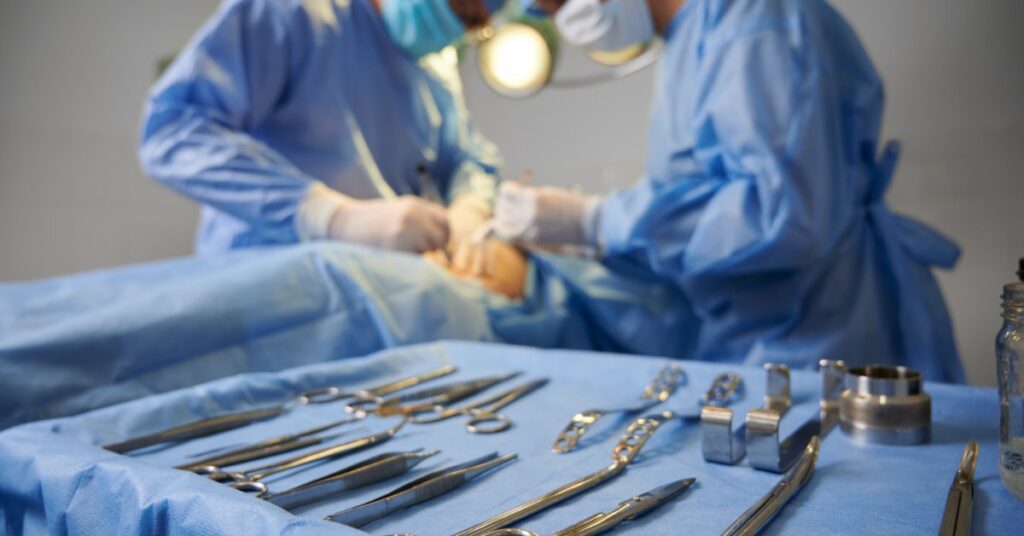 A tray of medical instruments rests on a blue sterile drape. Medical professionals perform surgery in the background.