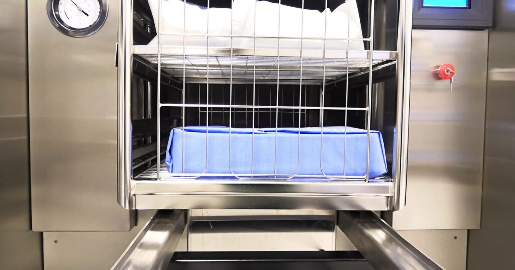 An open industrial steam sterilizer contains multiple shelves holding wrapped packs and a covered tray.