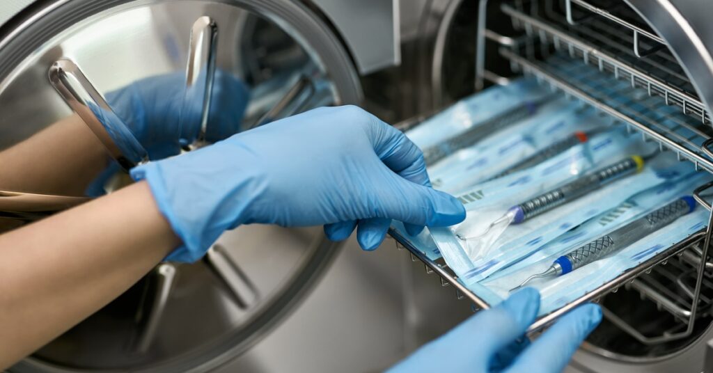 A person wearing blue gloves slides a tray of individually packaged dental instruments into a steam sterilizer.