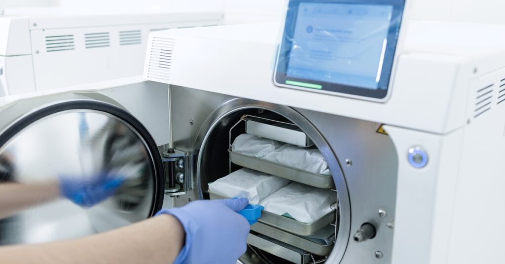 A person slides a metal tray of wrapped instrument packs into an autoclave. Several more trays are inside the chamber.