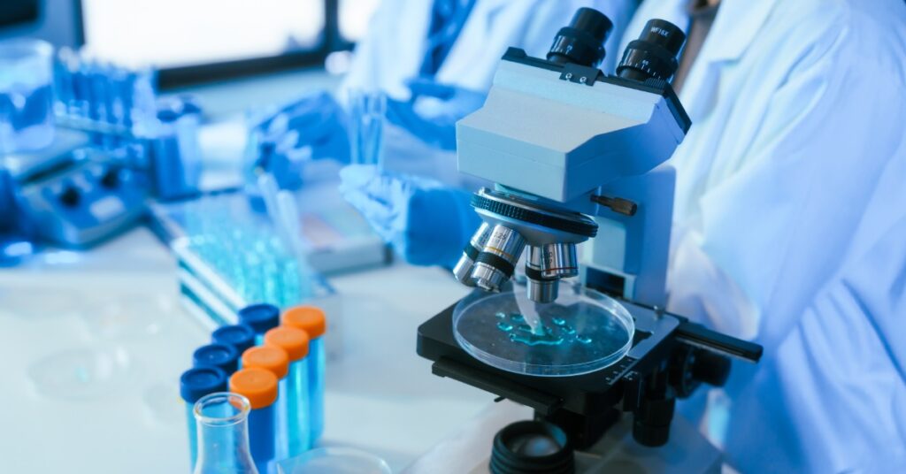 A laboratory microscope is over a petri dish with a blue liquid sample. More test tubes and lab workers are nearby.