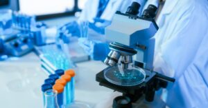 A laboratory microscope is over a petri dish with a blue liquid sample. More test tubes and lab workers are nearby.