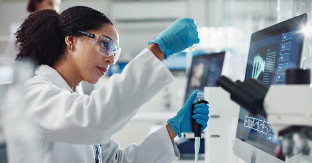 A scientist holds a sample container and a pipette. Computer monitors and a microscope are on the lab bench beside her.